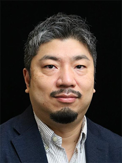 A professional headshot portrait of an unidentified male with short, gray hair, a goatee, and mustache, wearing a dark blazer and striped shirt, against a black background.