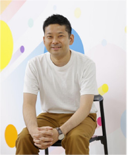 A man in a white t-shirt and brown pants sits and smiles in front of a colorful abstract background.