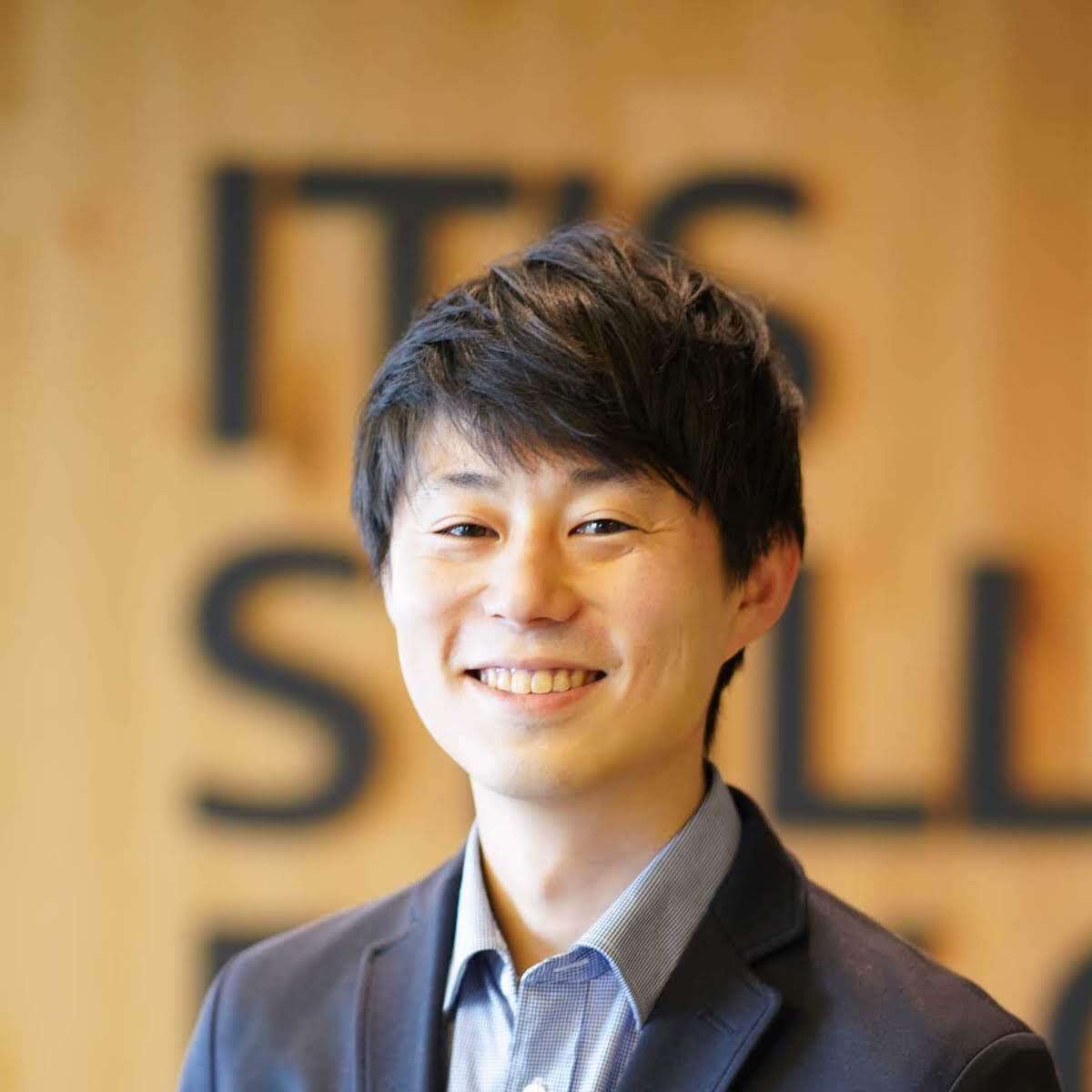 A professional headshot of a smiling individual in business attire, with a blurred background featuring large text on a wooden wall.