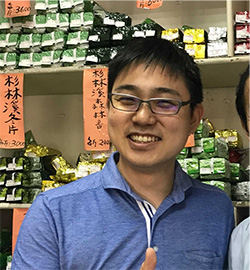 A person wearing glasses and a blue shirt is smiling inside a shop, with shelves filled with neatly stacked products and Japanese signage in the background.