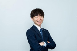 A business portrait of a young professional in a suit, standing with arms crossed and smiling against a plain background.
