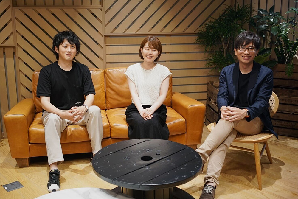 Three people are sitting and smiling in a modern office space with wooden panel walls, plants, and a round table. The atmosphere appears casual and friendly.
