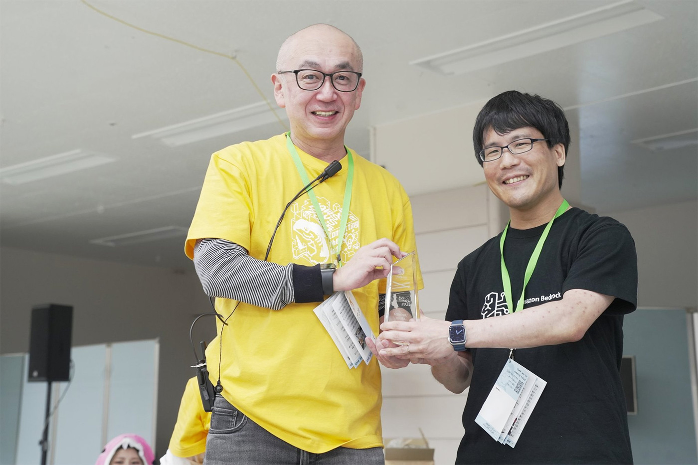 Two people smiling as one presents the other with an award at the AWS Samurai 2024 event. The presenters are wearing lanyards and event shirts, and the atmosphere is celebratory.