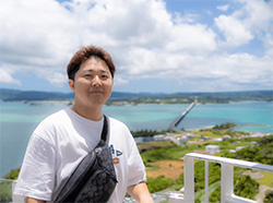 A person standing outdoors with a scenic view of the sea, a long bridge, and cloudy blue skies in the background.