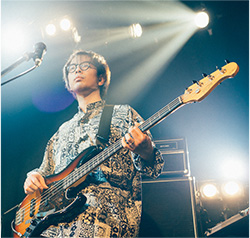 A musician is performing on stage, playing a bass guitar under stage lights with a microphone nearby.