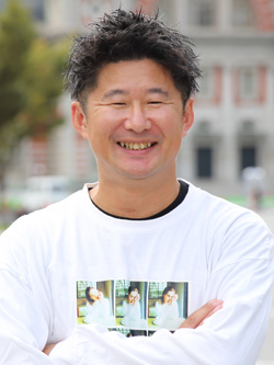 A man with short dark hair, wearing a white shirt with printed images, smiling outdoors with a blurred building in the background.