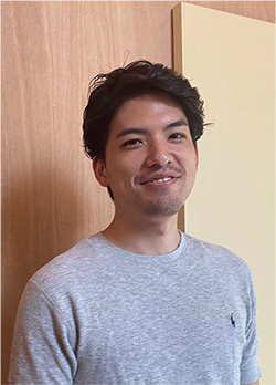 A casual portrait of a man with short black hair wearing a gray shirt, standing in front of a wooden and cream-colored background.