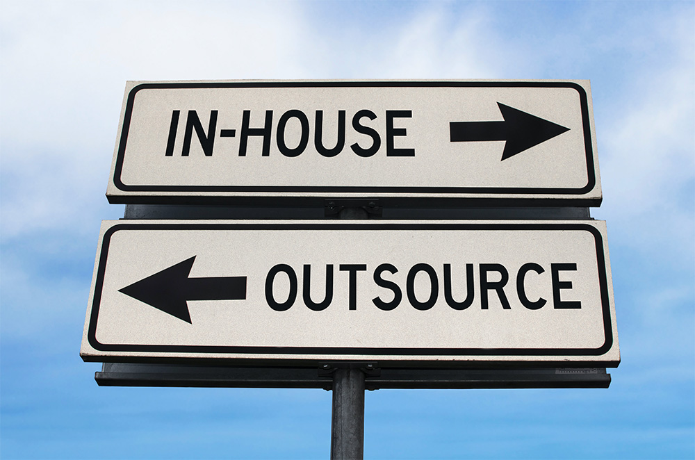 Two direction signs against the sky, one pointing right labeled 'IN-HOUSE' and one pointing left labeled 'OUTSOURCE', illustrating a choice between in-house and outsourcing options.