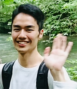 A person smiling and waving while standing outdoors with greenery and a river in the background.