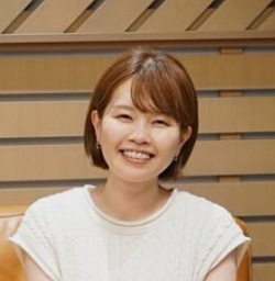 A woman smiling while seated indoors in a casual setting.