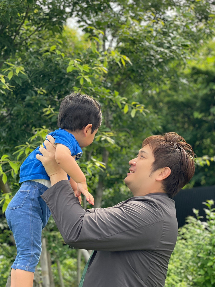 A father lifts his young son in the air while outdoors surrounded by green trees and foliage, expressing joy and family bonding in a natural setting.