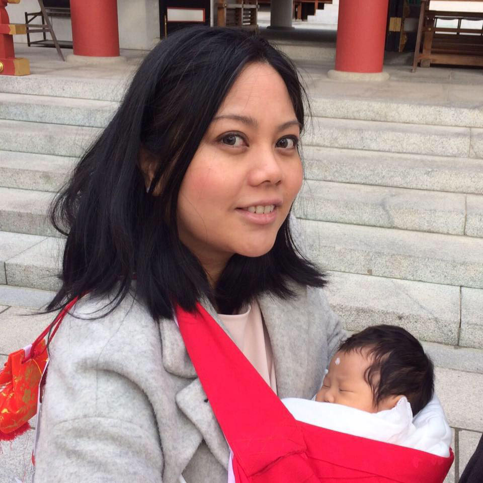 A woman holding a baby in a red sling while sitting on stone steps, with architectural columns in the background.