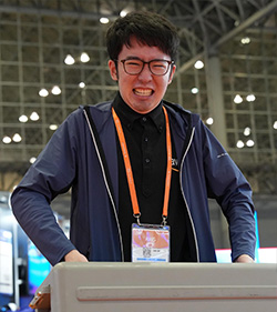 A person wearing glasses and an event badge is smiling while participating in an event. The background features a large indoor venue with spotlights and structural elements visible.