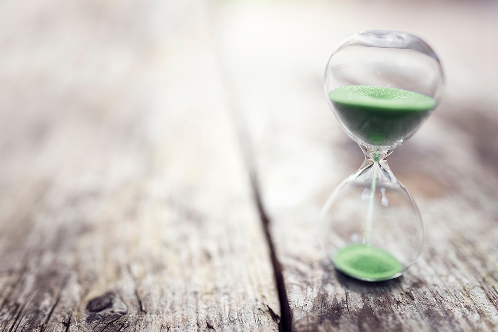 A glass hourglass filled with green sand sits on a rustic wooden table.