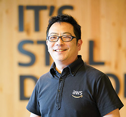 A portrait of an AWS employee wearing an AWS-branded shirt, standing in front of a wooden wall with large text.