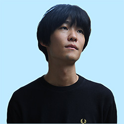 Portrait of an individual with dark hair wearing a black shirt, set against a light blue background.