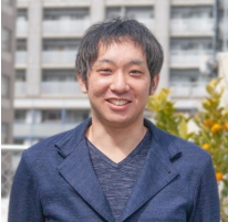 A man smiling outdoors with an urban background, featuring buildings and greenery.