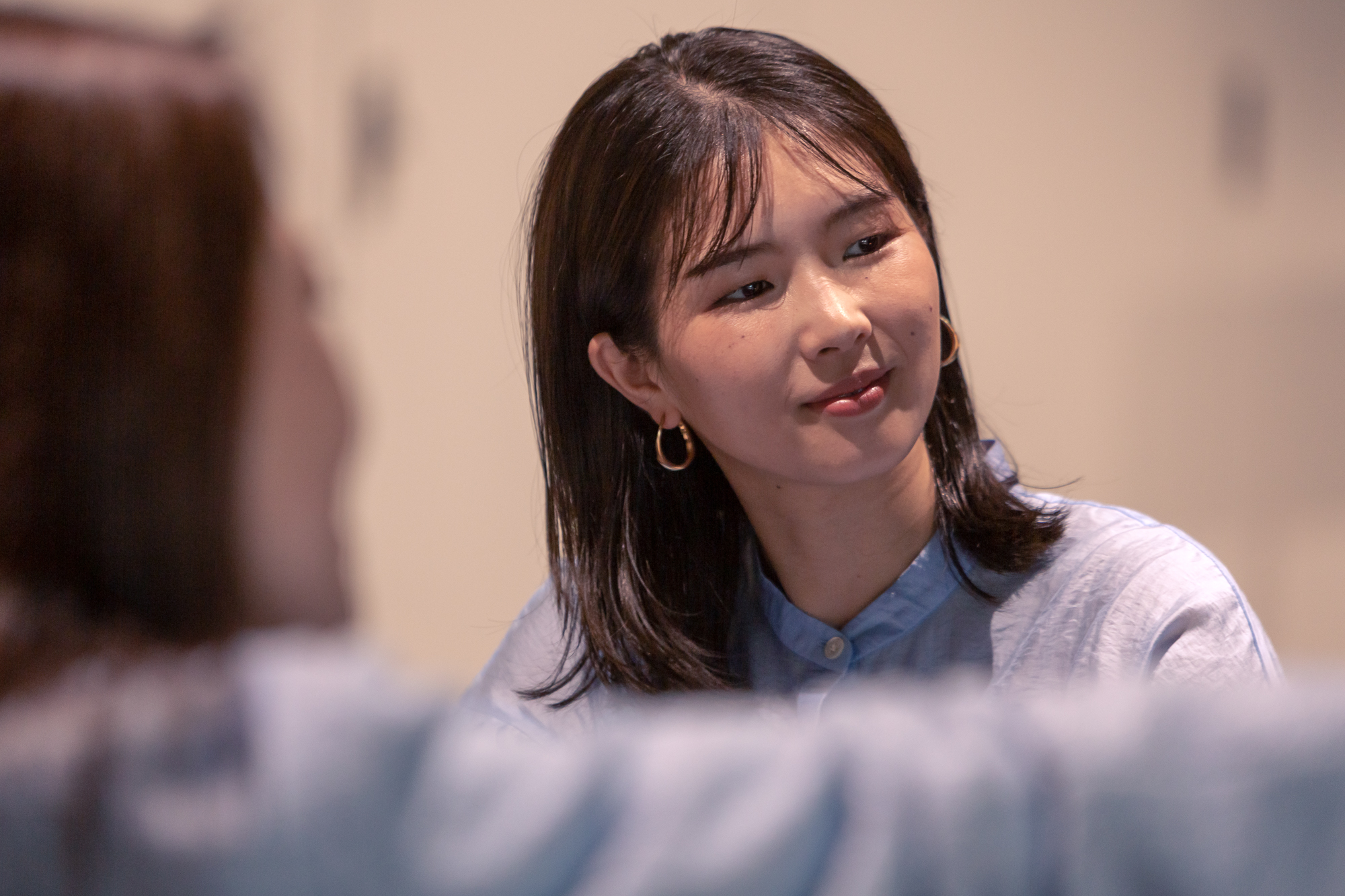 A woman is smiling and engaged in a casual conversation indoors, wearing a light blue shirt and gold hoop earrings.