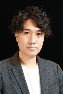 Formal portrait of a man with wavy hair, wearing a gray jacket and white shirt, against a black background.