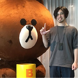 A man is standing and posing next to a large bear mascot figure indoors. The bear mascot has a round face with a neutral expression.
