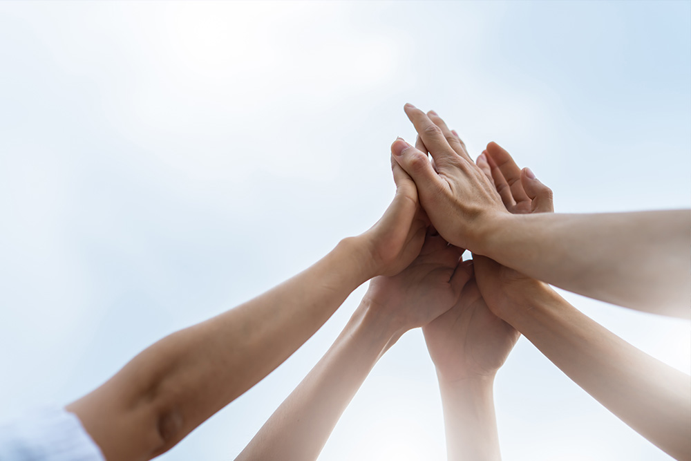 Multiple hands joining together for a high five against a bright sky, symbolizing teamwork and collaboration.