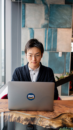 A person is working on a laptop with an AWS sticker in a contemporary office environment with patterned blue panels on the wall.