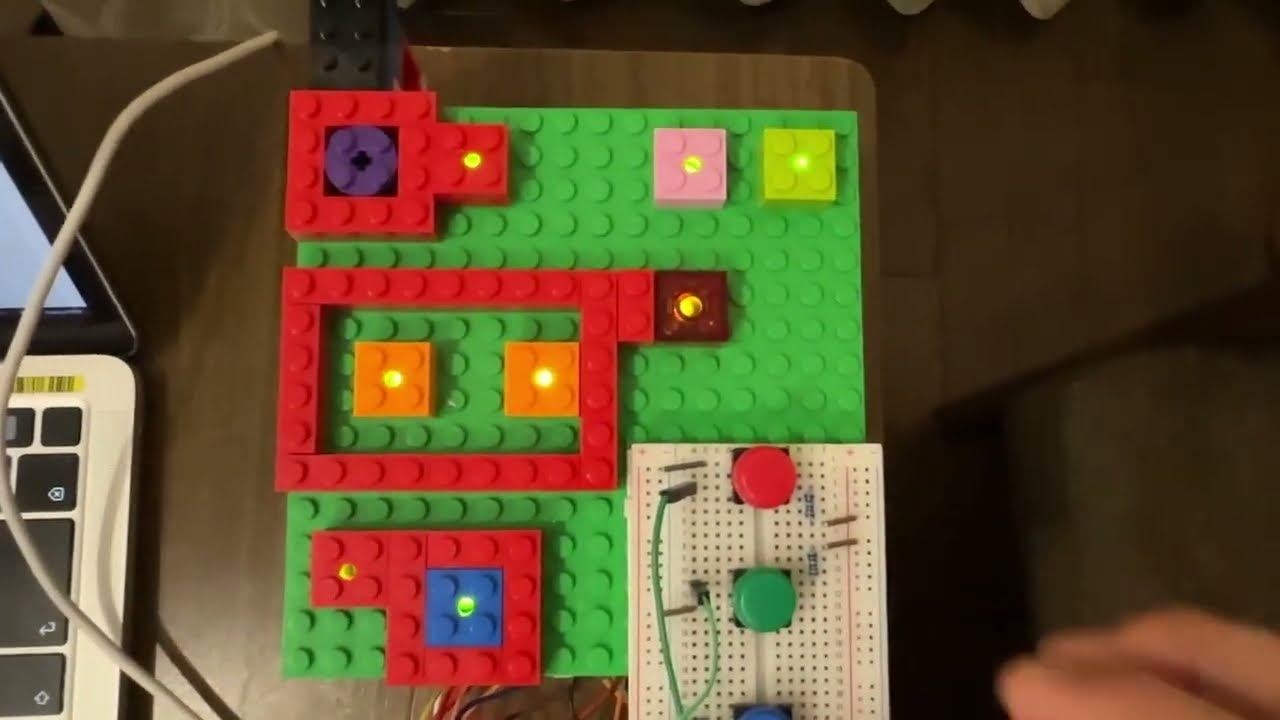 A top-down view of a LEGO baseplate with colorful bricks arranged as a circuit board, featuring illuminated LEDs and a small breadboard with buttons, next to a laptop.