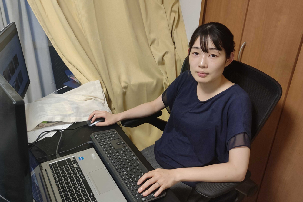 A woman sits at a desk working on a laptop with an external keyboard and mouse in a home office environment.