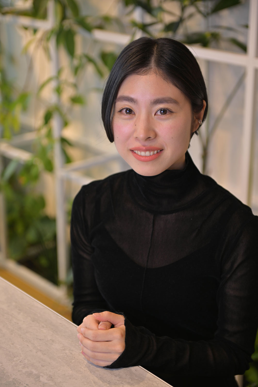 A woman wearing a black top is sitting at a table with her hands folded. Indoor green plants are visible in the background.