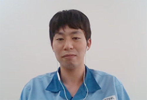A man wearing a blue shirt and earphones, seated against a plain background.