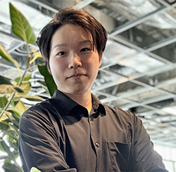 A professional portrait of a person wearing a dark shirt, standing in an office environment with industrial-style ceilings and green plants in the background.