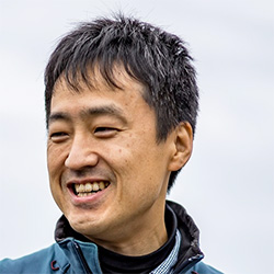 A portrait of a smiling man outdoors, wearing a teal jacket, with a blurred sky background.