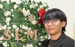 A person with glasses and dark hair is standing in front of a floral arrangement with the letters 'SEM' and a red ribbon decoration.