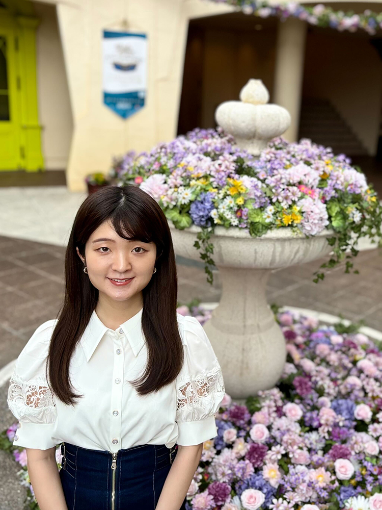 A woman in a white blouse standing in front of a decorative fountain surrounded by colorful flowers.