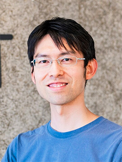 A portrait photograph of a person with short black hair, wearing glasses and a blue shirt, smiling in front of a neutral textured background.