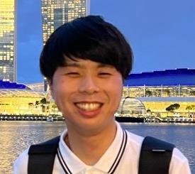 A man smiling in front of a waterfront cityscape during the evening, with illuminated modern buildings in the background.
