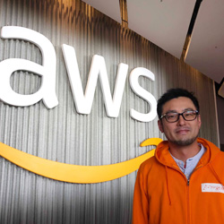 A person wearing an orange jacket stands in front of an AWS logo sign inside an office setting.
