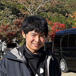 A person smiling outdoors in a park-like setting with colorful trees in the background, wearing a jacket. Photo taken in 2023.