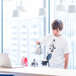 A person pours hot water from a kettle into a coffee dripper in a modern office with large windows, wearing a graphic t-shirt and standing near a laptop.