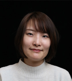 Portrait photo of a woman with short brown hair wearing a white sweater, set against a black background.