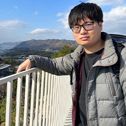 A person wearing glasses and a gray jacket poses outdoors on a bridge with mountains and a blue sky in the background.