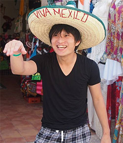 A man smiling and wearing a large sombrero with 'Viva Mexico' written on it, standing in a colorful market.