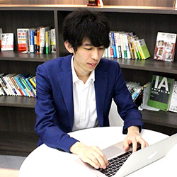 A person in a blue jacket is working on a laptop at a white desk, with a bookshelf filled with books in the background.