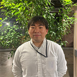 A person wearing a white shirt standing indoors in front of large green indoor plants.