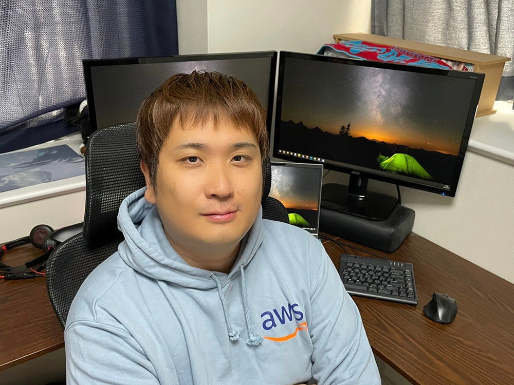 A person wearing an AWS hoodie sits at a desk with multiple computer monitors in the background.