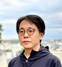 A portrait of a man with short black hair, wearing round glasses and a dark jacket, standing outdoors with a blurred cityscape background.