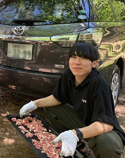 A person kneels outdoors holding a tray of raw meat, preparing for outdoor cooking beside a Toyota car.