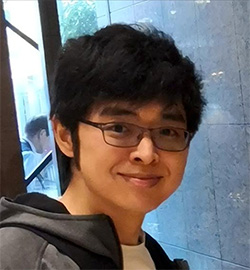 A portrait photo of a person with short dark hair and glasses, smiling and wearing a jacket, standing indoors in front of a reflective wall.