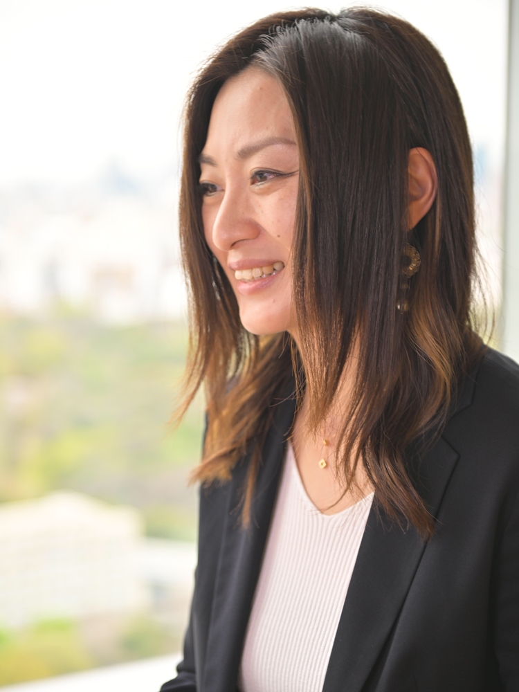 A woman wearing business attire smiles while standing near a window with daylight illuminating her face.