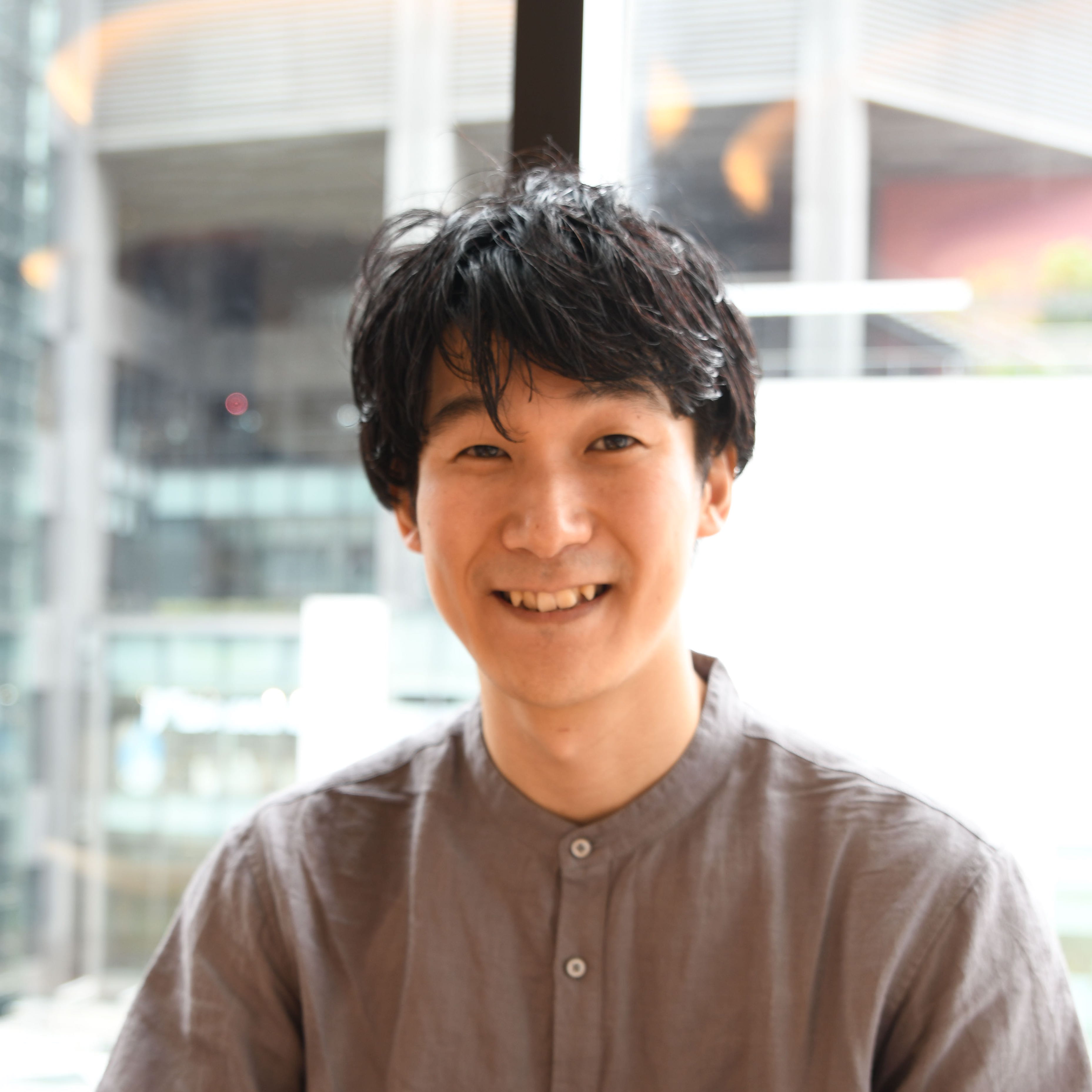 A man in a casual shirt smiling indoors with natural light coming through the window in the background.